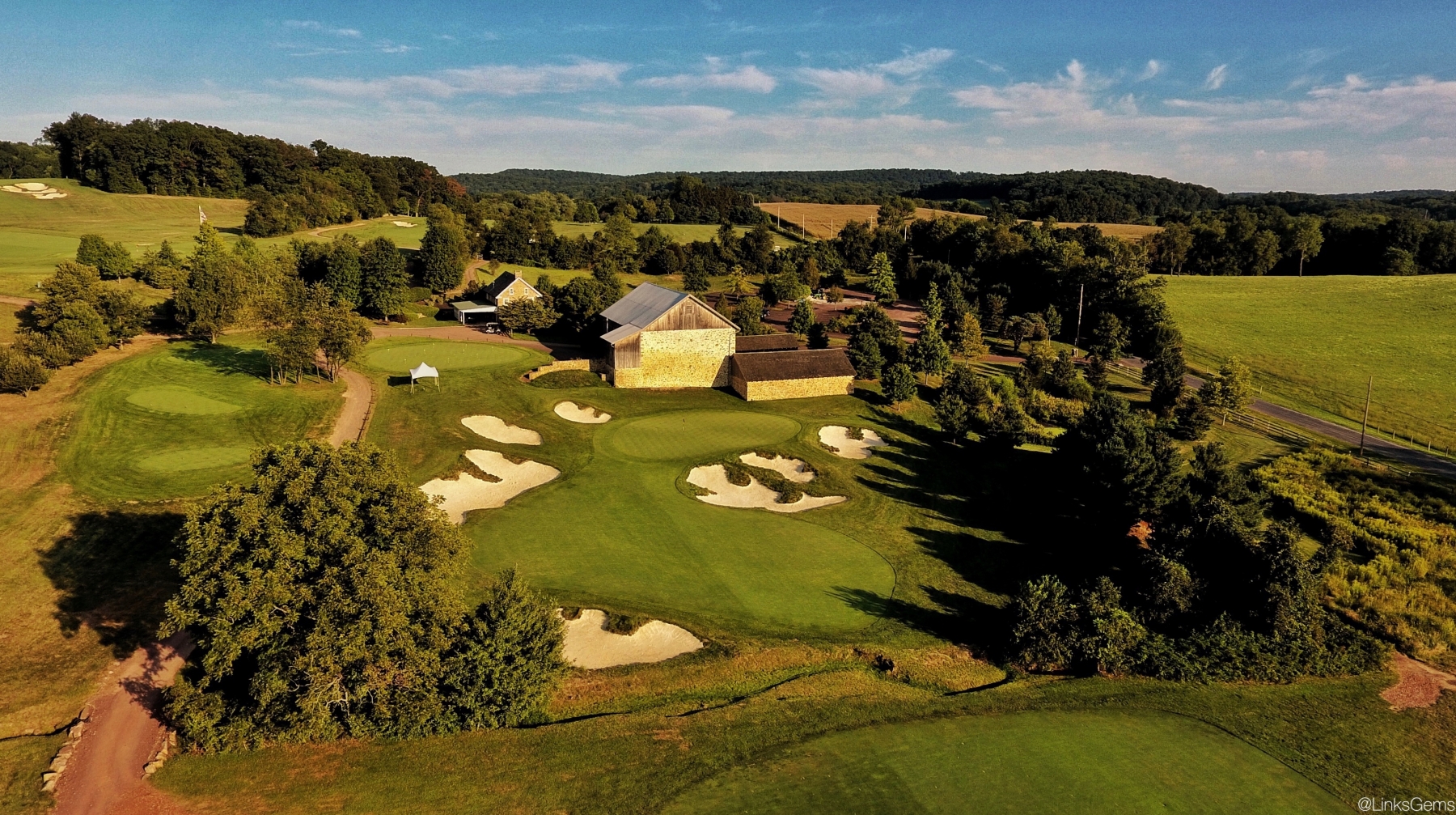 Aerial view of a golf course surrounded by rolling countryside, featuring a practice green and multiple white sand bunkers arranged around a central putting area. A large rustic barn and clubhouse sit beside the course, while tree-lined fairways, open fields, and winding paths extend across the landscape under a clear blue sky.