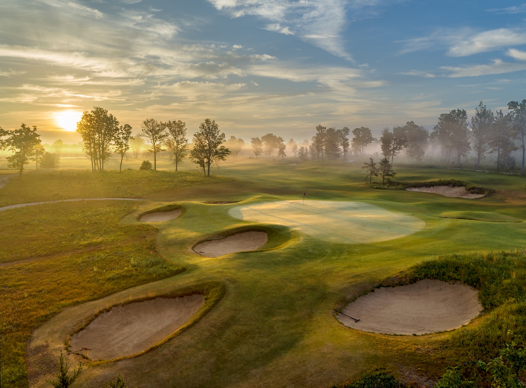 Aerial view of a golf course at sunrise, featuring a large green surrounded by sculpted sand bunkers. Golden morning light and low-lying mist stretch across the fairway, while trees line the horizon beneath a sky filled with soft clouds. The scene highlights the dramatic contours of the course and the tranquil atmosphere of an early morning round.
